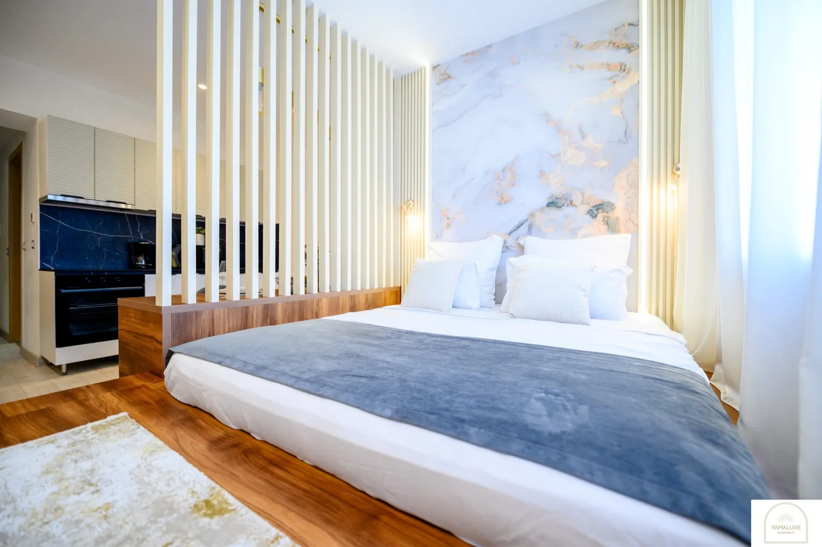 Low wooden platform bed against a backlit marble-effect feature wall, with cream slat divider and kitchenette behind (5/5)