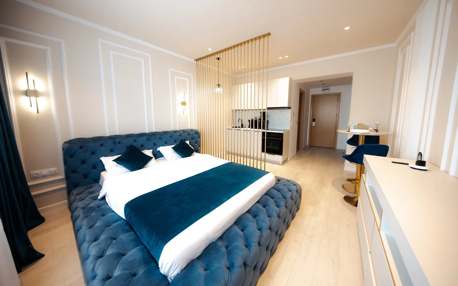 Blue tufted velvet bed in an elegant studio with gold rod room divider, white kitchenette and marble bistro table (1/5)