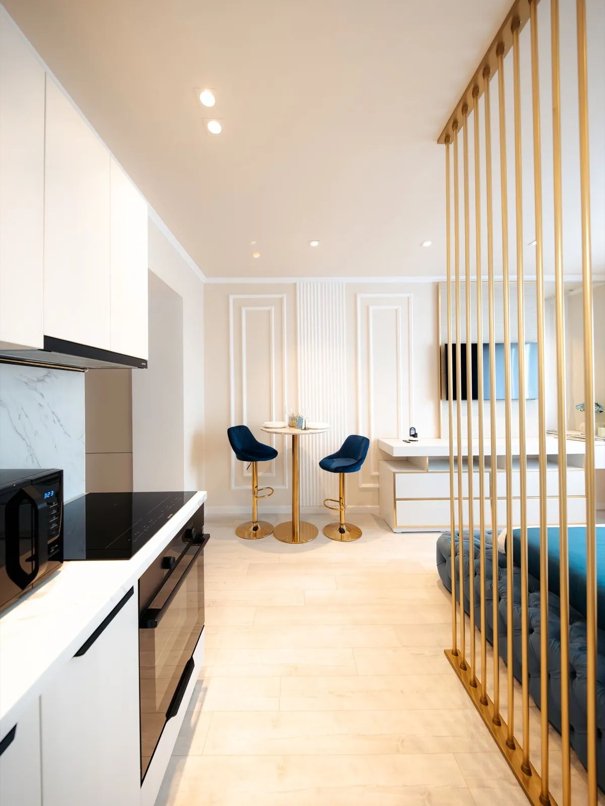 White kitchen units with microwave and oven, leading to a marble bistro table with navy velvet bar stools on gold legs (2/5)