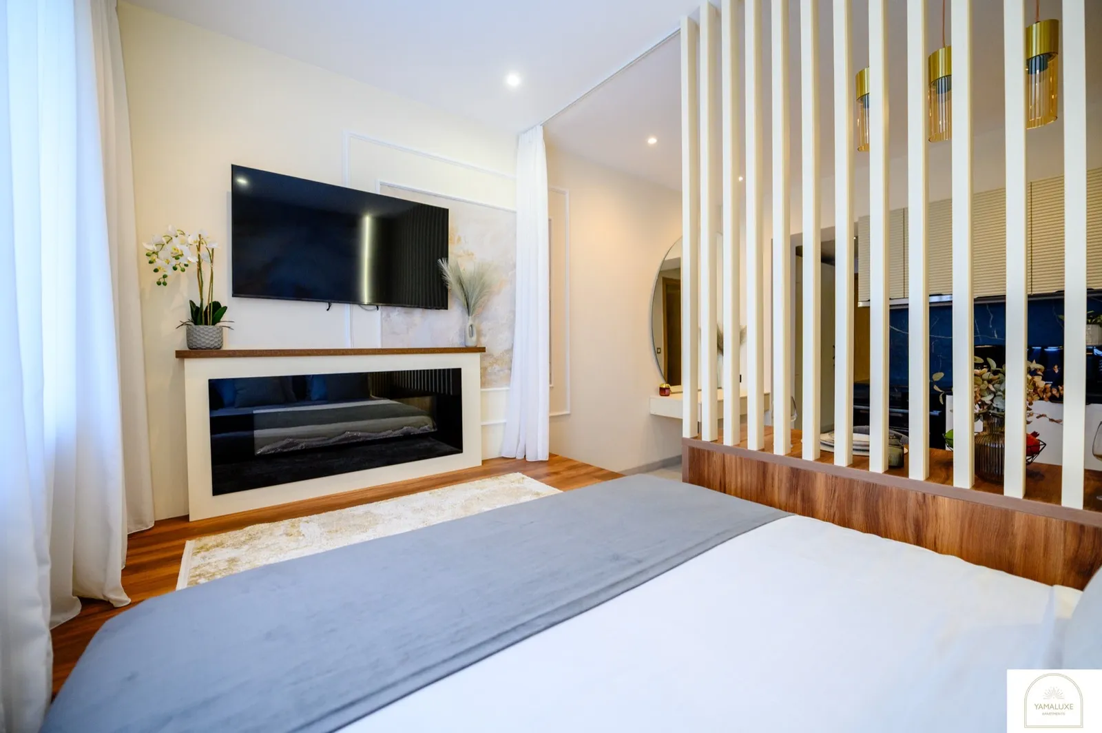Wall-mounted TV above a mantel-style console facing the bed, with round vanity mirror, orchids and cream slatted partition (3/5)