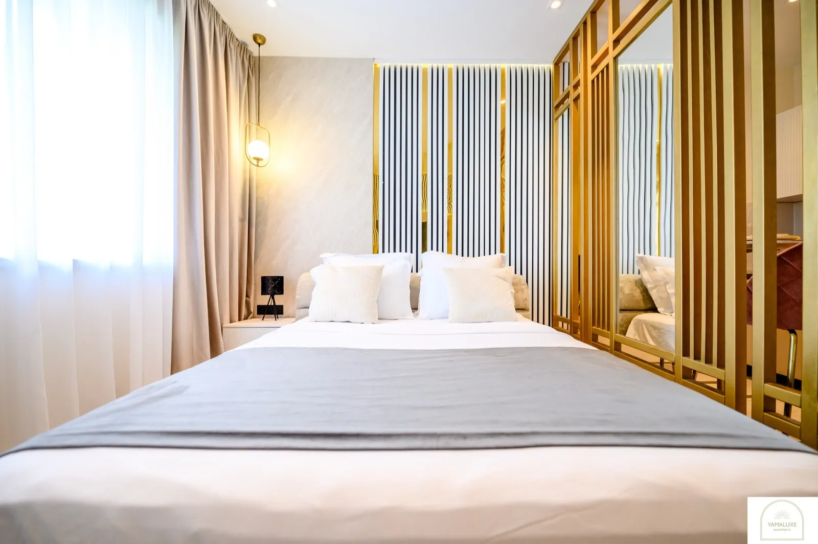 Double bed with plush white pillows against a backlit black, white and gold striped wall, with mirrored gold panels (4/5)