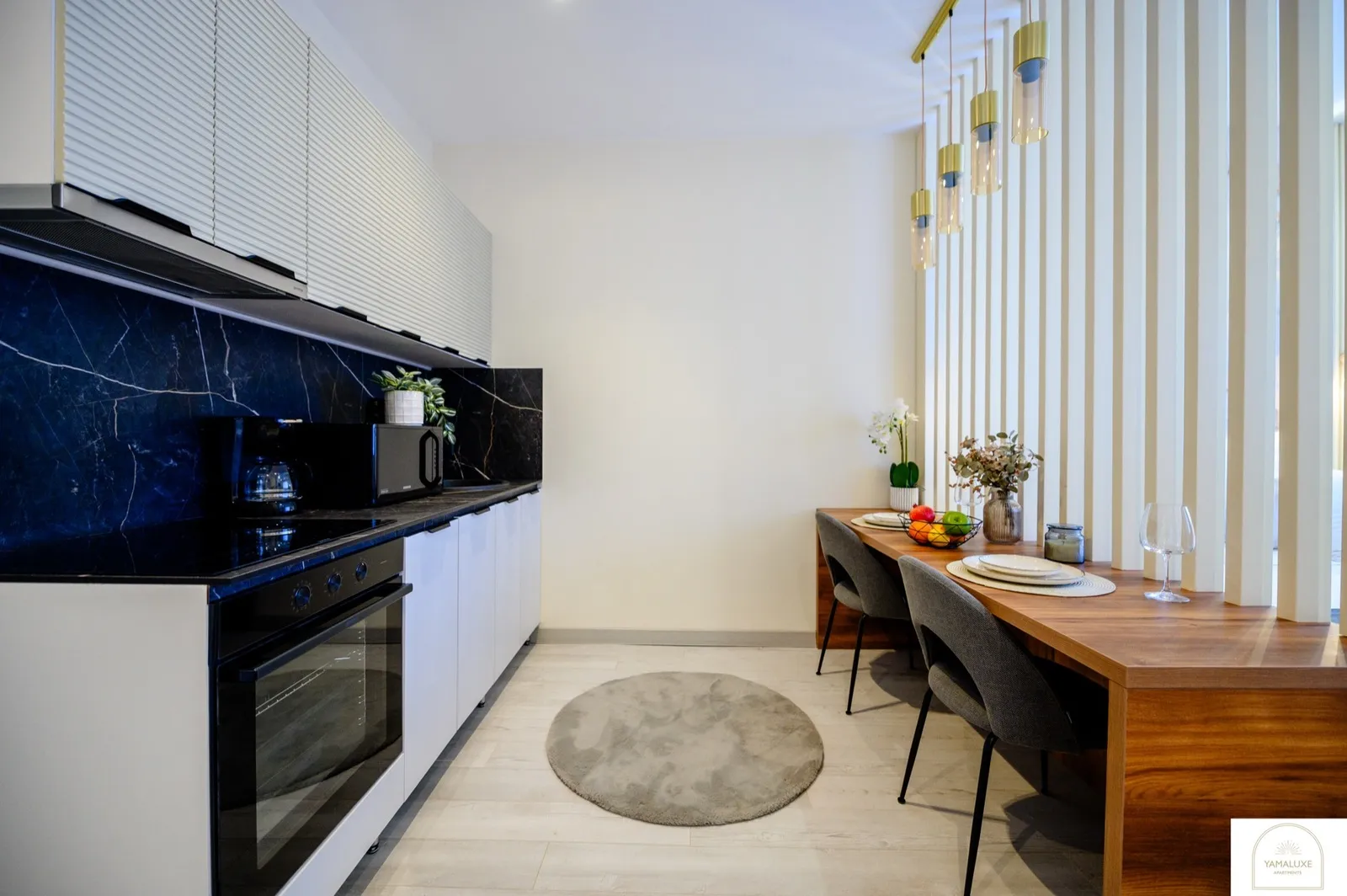 Bucătărie cu backsplash din marmură bleumarin, dulapuri albe striate, cuptor negru și masă din lemn sub pendule aurii