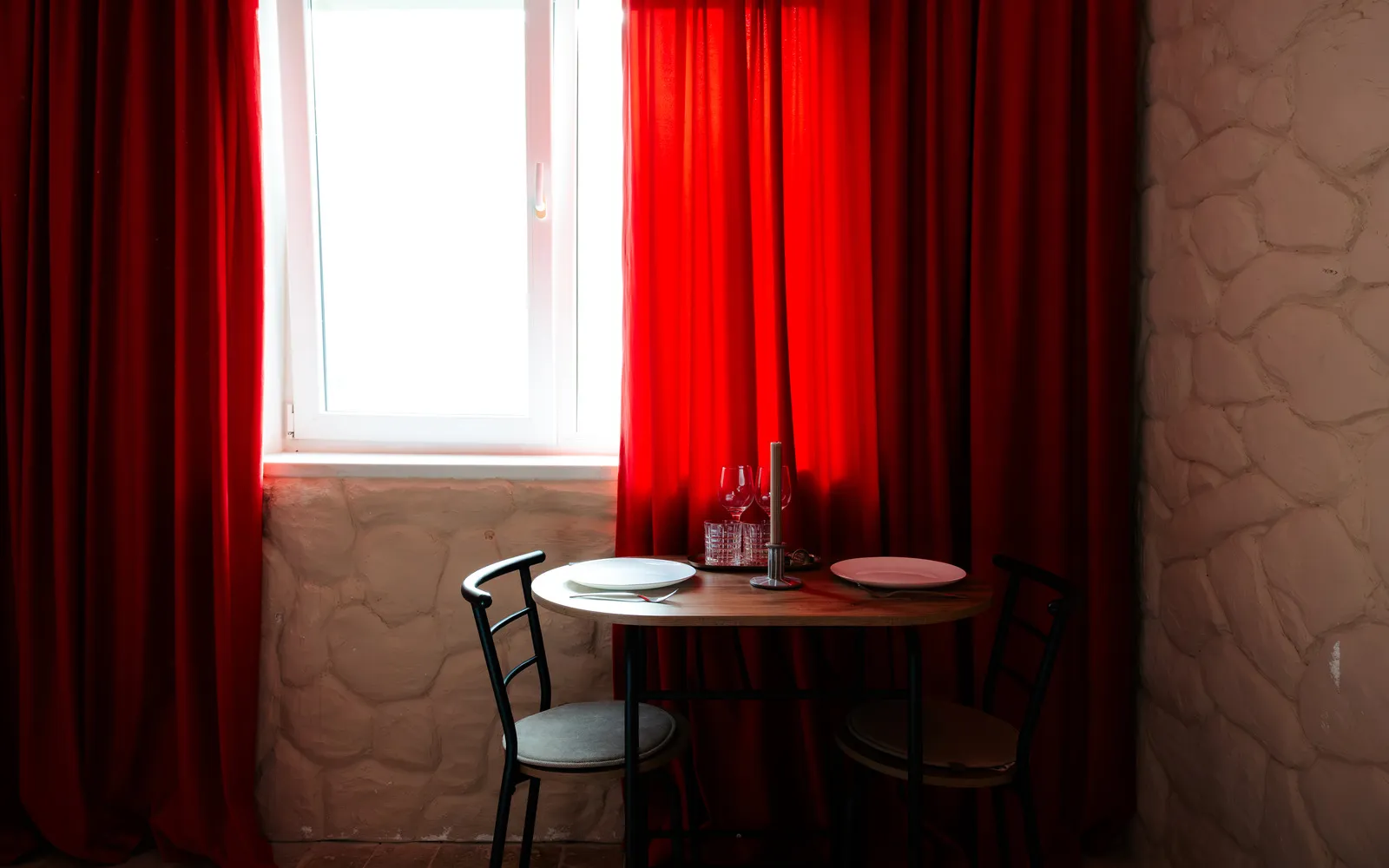 Masă de două persoane lângă fereastră, cu pahare de vin, pahare de apă și o lumânare, între draperii roșii de catifea