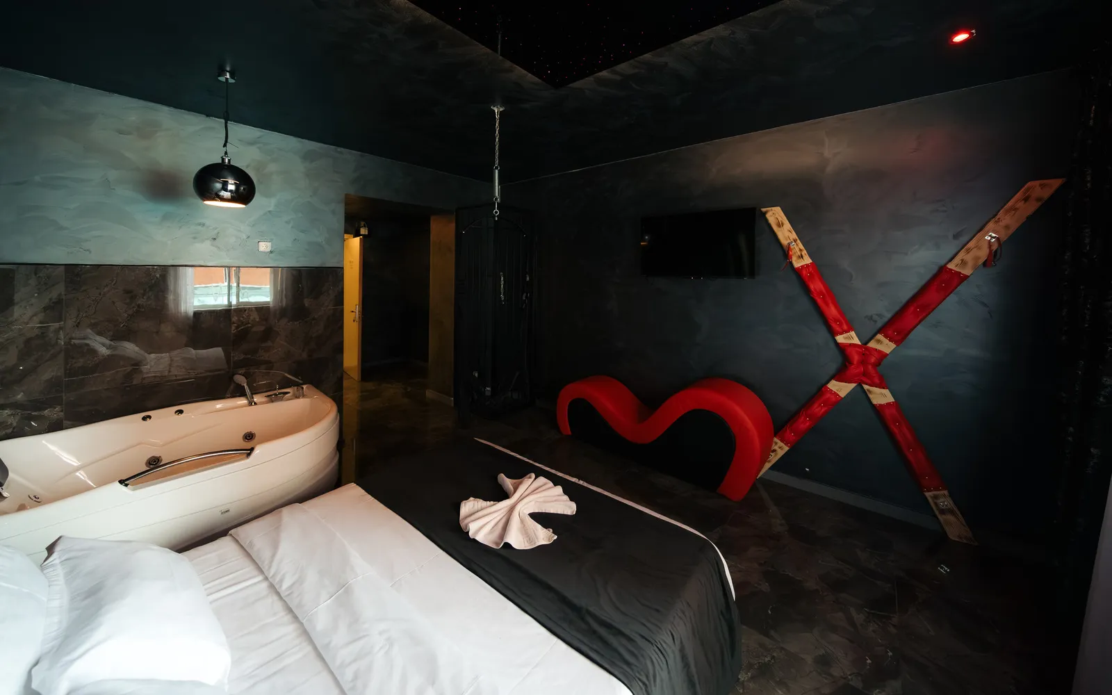 Cameră în tonuri închise, cu cadă cu hidromasaj pe colț, șezlong tantra roșu, cruce cu sistem de fixare pe perete și tavan înstelat cu fibră optică