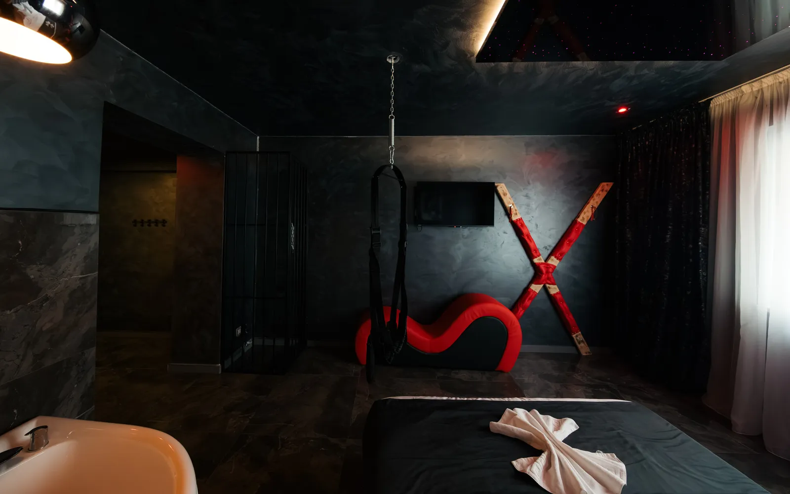 Șezlong tantra roșu și cruce montată pe perete lângă un leagăn suspendat din tavan, cu cada cu hidromasaj în prim-plan