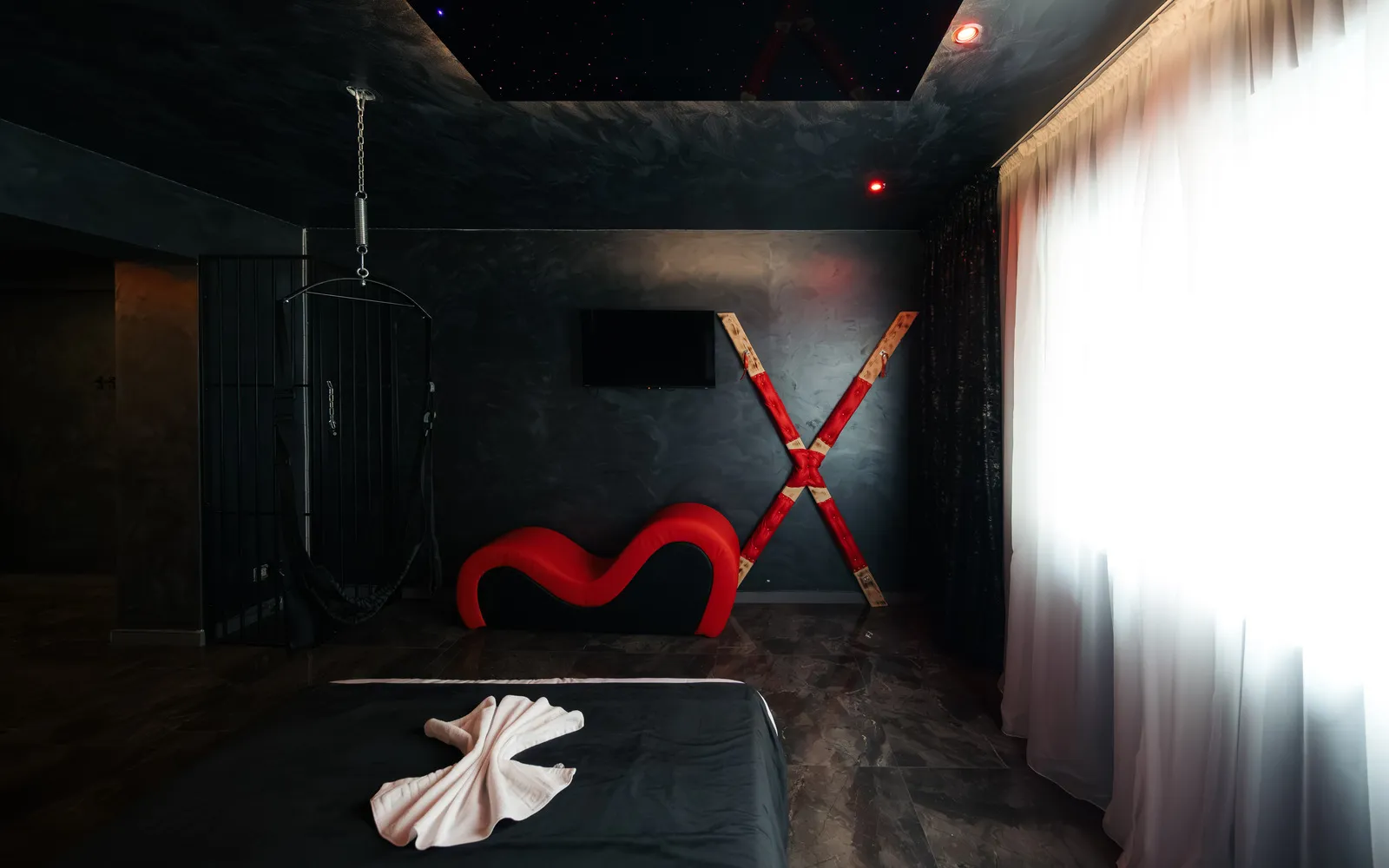 Șezlong tantra roșu și cruce cu puncte de fixare, pe pereți în tencuială închisă la culoare, leagăn și incintă cu gratii în stânga